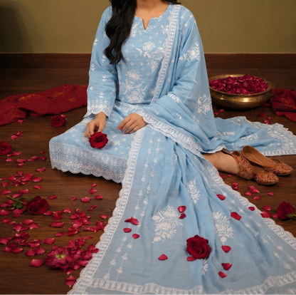A woman sits on a wooden floor wearing a light blue embroidered traditional outfit with a matching dupatta. The outfit features intricate white floral embroidery and border details.